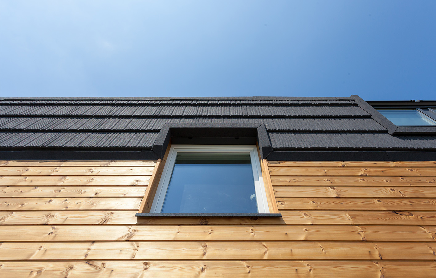 Moderne Holzfassade mit Dachabschluss – Blick auf Qualität und Design Nahaufnahme einer modernen Holzfassade mit Fenster und schwarzem Dachabschluss bei strahlend blauem Himmel.